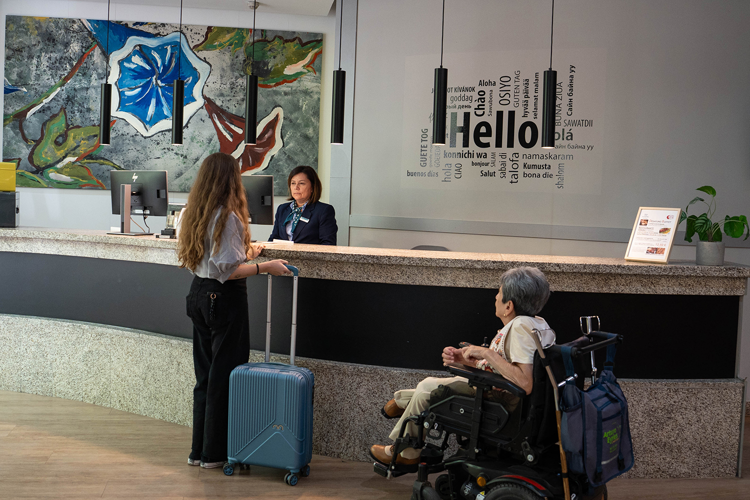 Una mujer en silla de ruedas llega a la recepción de un hotel acompañada por su asistente personal que lleva una maleta.