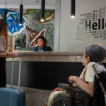 Una mujer en silla de ruedas delante de la recepción de un hotel acompañada de su asistente personal llevando una maleta y hablando con la recepcionista.