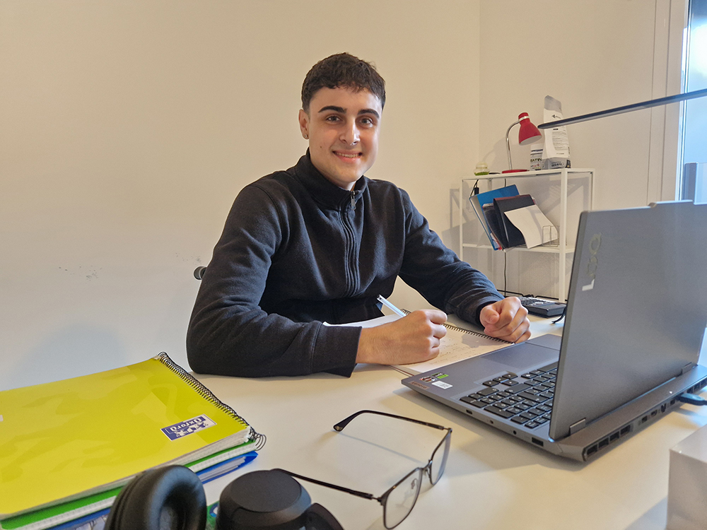Eloy en su escritorio estudiando con el portátil, mirando a cámara sonriente.