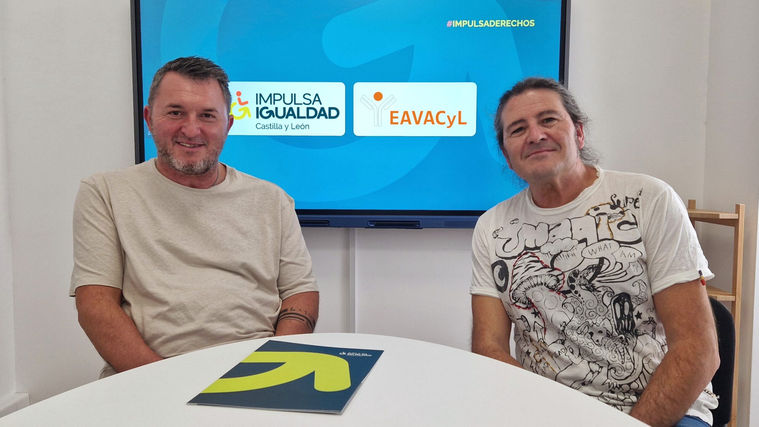Posan mirando a cámara, a la izquierda, Francisco Sardón, y a la derecha, José Andrés Herranz. De fondo, en la pantalla, los logos de IMPULSA IGUALDAD Castilla y León y EAVACyL y el hashtag IMPULSADERECHOS. En primer término, una mensa con una carpetilla encima de IMPULSA IGUALDAD Castilla y León.