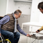 En un salón, un asistente personal enciende un reproductor de CD con un mando a distancia, mientras un usuario en silla de ruedas ajusta una de las ruletas del aparato. sirve un vaso de agua al usuario en silla de ruedas.