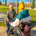 Fotografía en un parque soleado donde dos hombres jóvenes conversan alegremente. En primer plano y de espaldas se encuentra una persona en silla de ruedas, usando un gorro amarillo, una bufanda a juego y una chaqueta acolchada verde y blanca. En frente, sentado en un banco de madera, su compañero sonríe mientras lo escucha; viste una chaqueta verde oliva, un gorro rosa y una bufanda gris. Alrededor hay césped verde y árboles