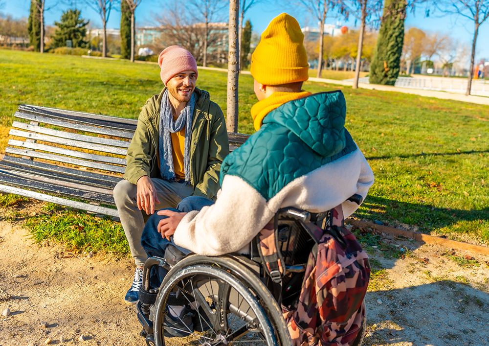 Fotografía en un parque soleado donde dos hombres jóvenes conversan alegremente. En primer plano y de espaldas se encuentra una persona en silla de ruedas, usando un gorro amarillo, una bufanda a juego y una chaqueta acolchada verde y blanca. En frente, sentado en un banco de madera, su compañero sonríe mientras lo escucha; viste una chaqueta verde oliva, un gorro rosa y una bufanda gris. Alrededor hay césped verde y árboles