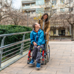 De paseo por la acera, un chico en silla de ruedas mirando hacia atrás a su asistente personal, ambos sonriendo.