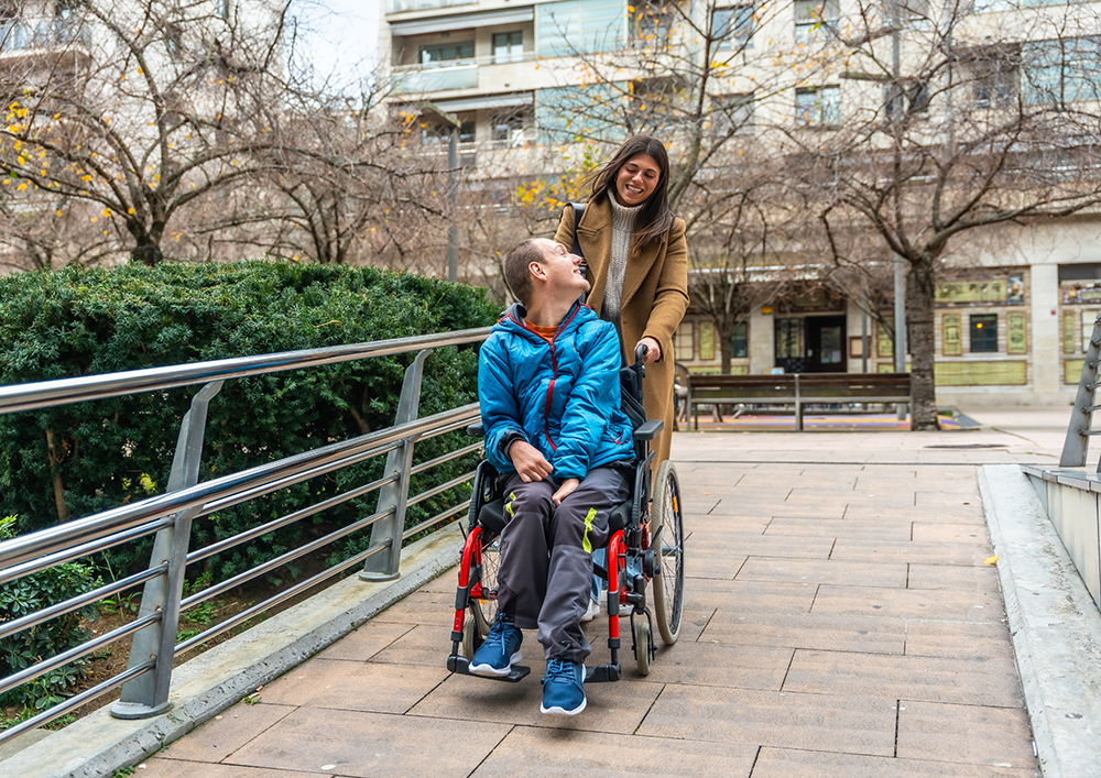De paseo por la acera, un chico en silla de ruedas mirando hacia atrás a su asistente personal, ambos sonriendo.